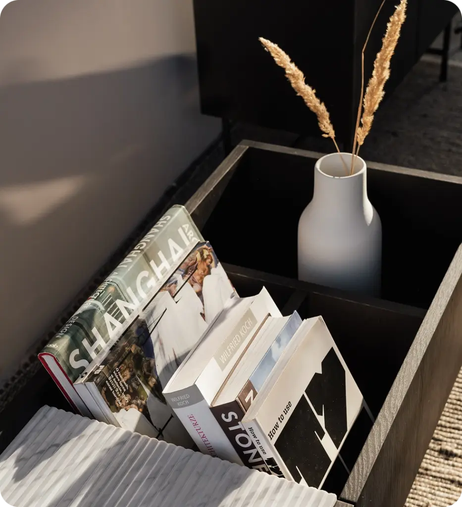 Eine schwarze Holzkiste mit Büchern zu verschiedenen Themen und einer weißen Vase mit Trockenblumen